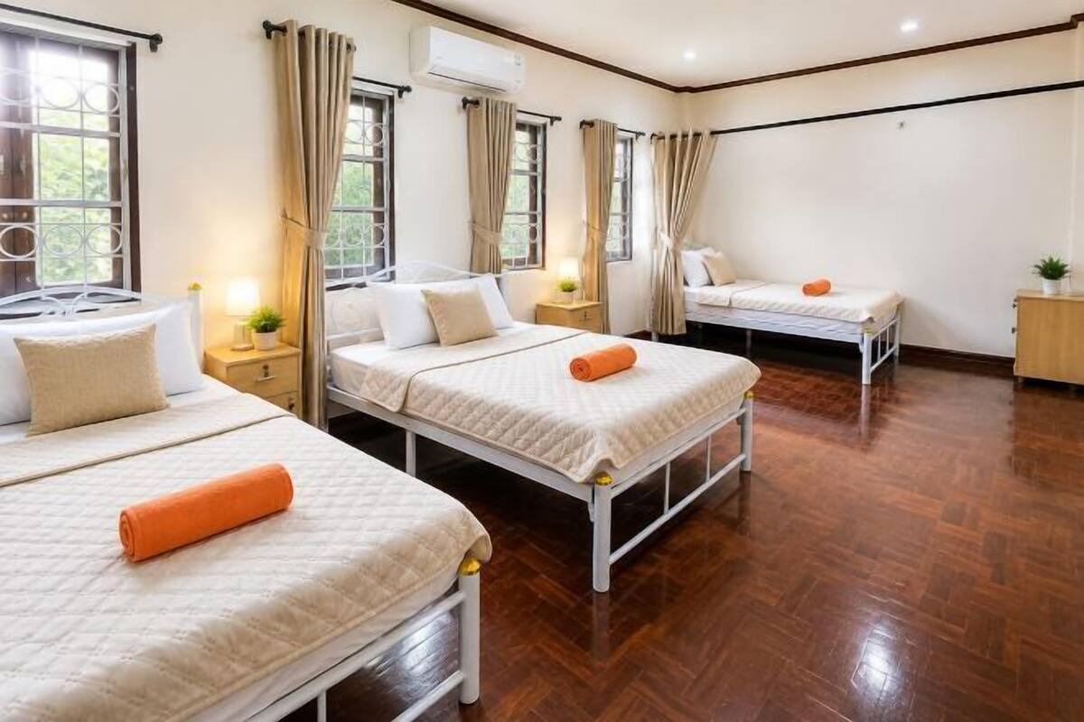 A hotel room with three beds, wooden flooring, and large windows with curtains.