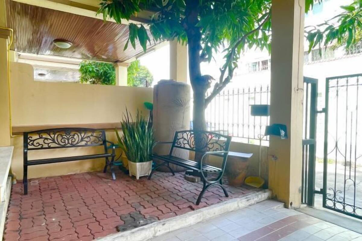 A covered outdoor seating area with a bench, potted plants, and a tree.