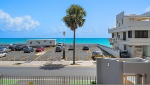 A parking lot with several cars, a palm tree, and a building with a balcony overlooking the ocean. - Coastal Charm Panoramic Scenes Direct Shorelin (San Juan)