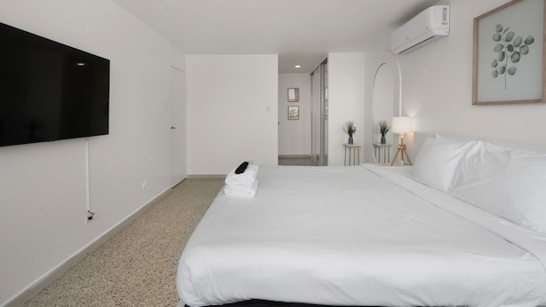A hotel room with a large bed, a flat-screen TV mounted on the wall, a bedside table with a lamp, and an air conditioning unit. - Coastal Charm Panoramic Scenes Direct Shorelin (San Juan)