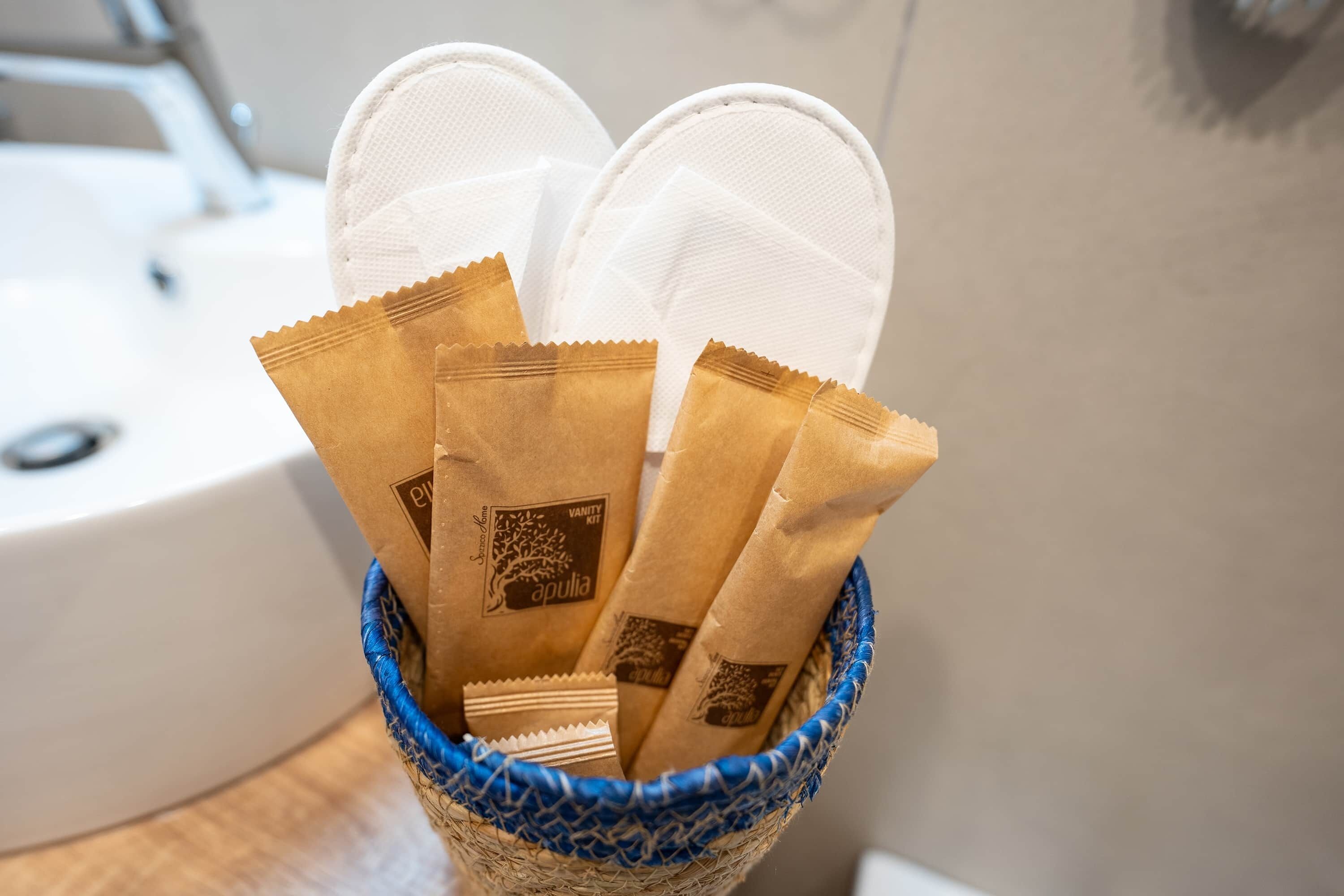 Una cesta con varias bolsas de papel marrón que tienen un logotipo, colocada sobre una superficie cerca de un lavabo blanco.