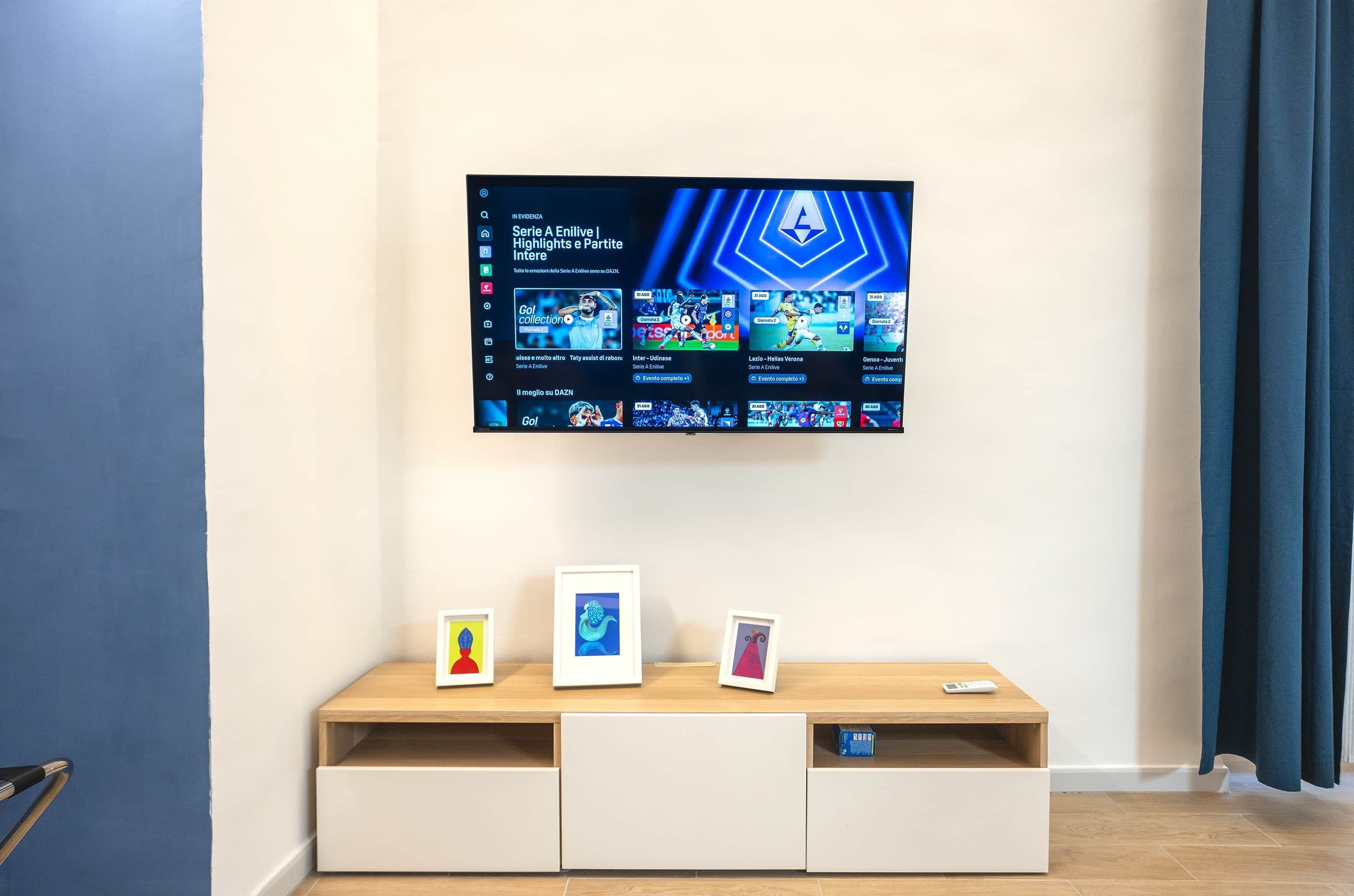 A modern living room with a flat-screen TV mounted on the wall displaying a video game interface. There is a wooden console table with three framed pictures and a remote control.