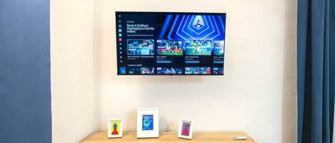 A modern living room with a flat-screen TV mounted on the wall displaying a video game interface. There is a wooden console table with three framed pictures and a remote control.