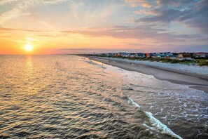Uma praia com um pôr do sol, águas calmas e casas na orla.