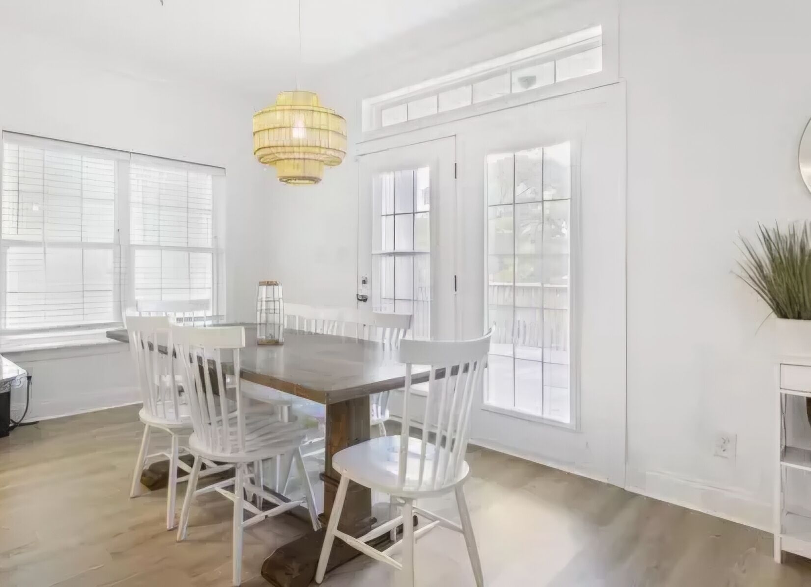 A dining area with a wooden table and white chairs, large windows, and a hanging light fixture.