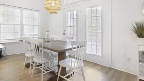 Un espace repas avec une table en bois et des chaises blanches, de grandes fenêtres et un luminaire suspendu.