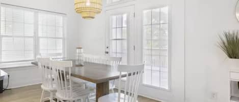 A dining area with a wooden table and white chairs, large windows, and a hanging light fixture.