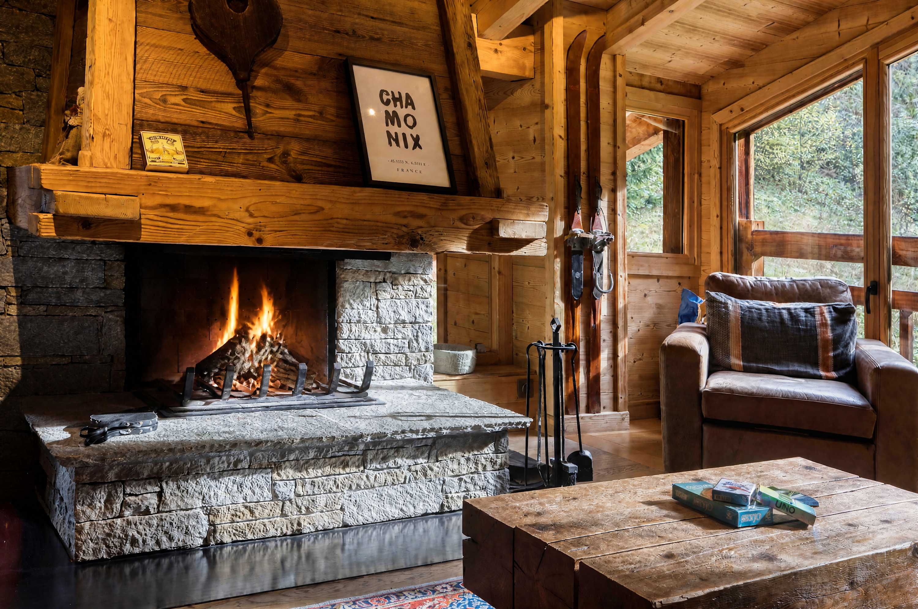 L’intérieur chaleureux d’un chalet avec une cheminée en pierre, des murs en bois, un coin salon confortable et une fenêtre donnant sur le paysage extérieur.