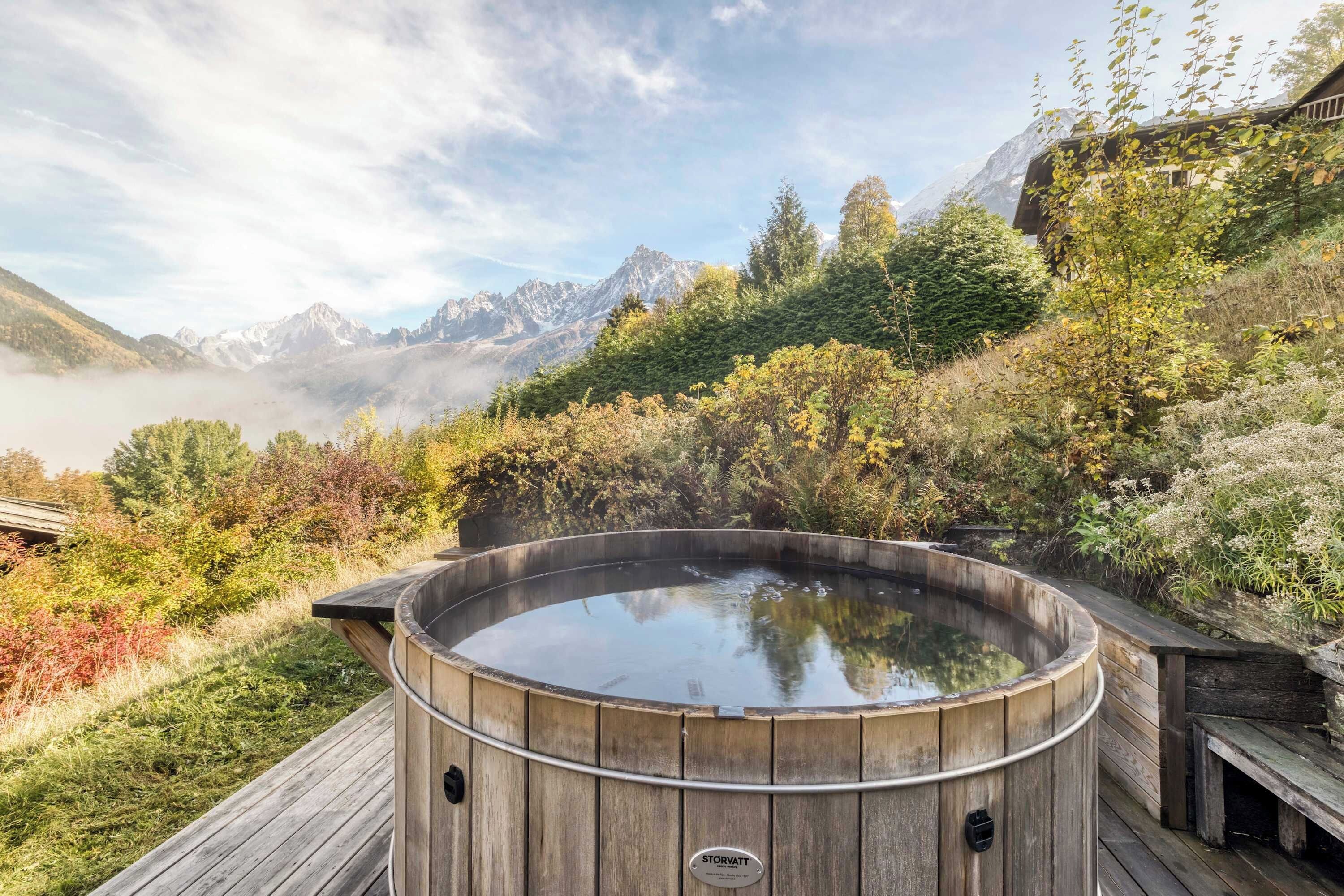A wooden hot tub on a deck with a mountainous backdrop.