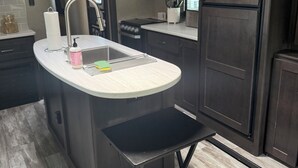 A modern kitchen with dark wood cabinets, a stainless steel refrigerator, a microwave, and a sink with a bar stool. - Cozy RV in Cool Columbus with AC (Columbus)