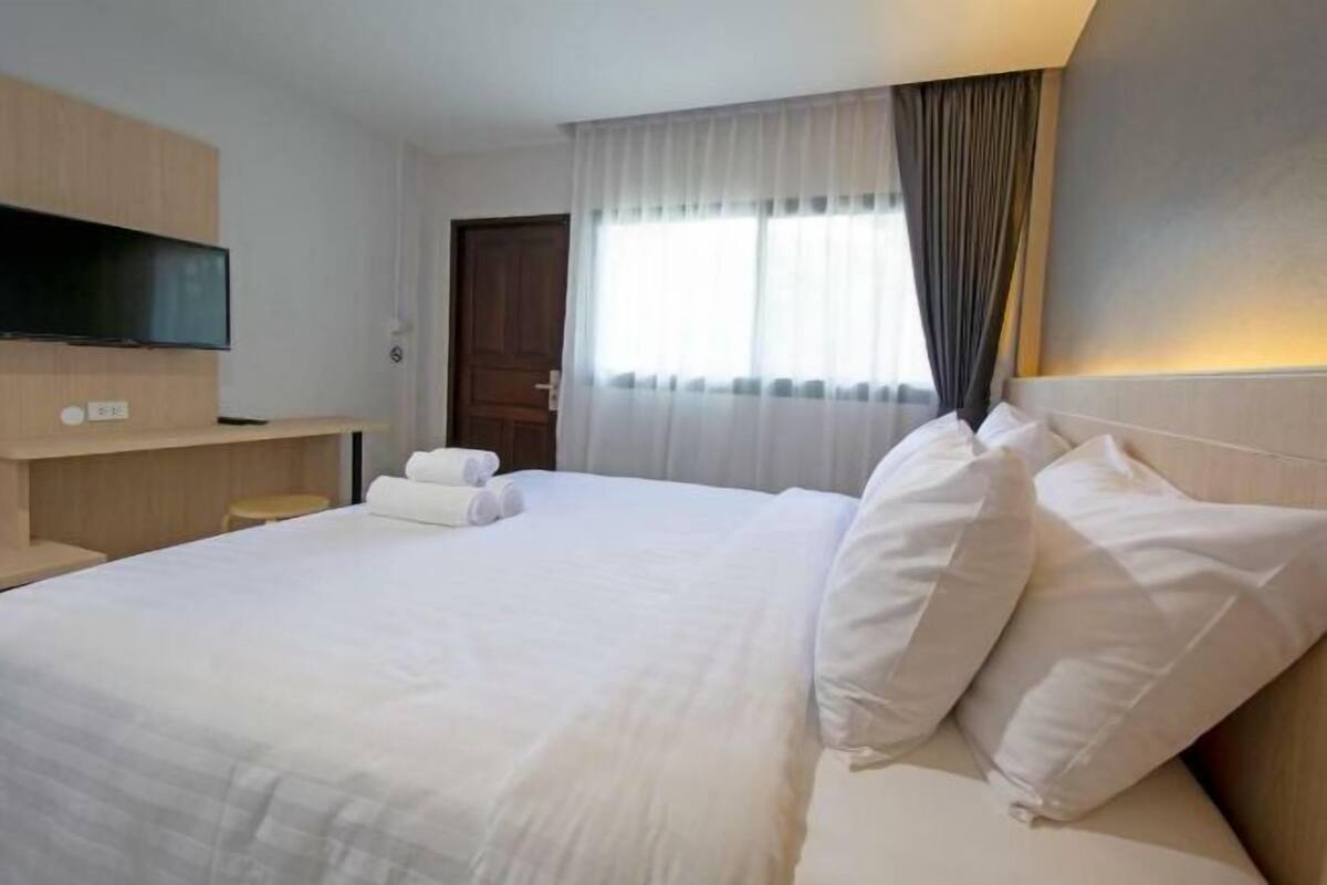 A hotel room with a large bed, a flat-screen TV mounted on the wall, a bedside table, and a window with sheer curtains.