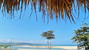 A beach view with a stone wall, a thatched roof, and a clear blue sky. - Three Queen Villa and Homestay (Wanokaka)