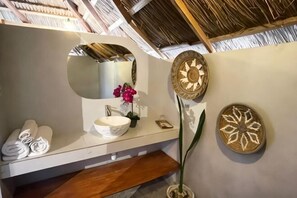 A bathroom with a round mirror, a sink, a towel rack, decorative wall art, and a plant. - Three Queen Villa and Homestay (Wanokaka)