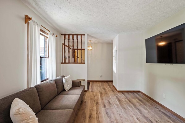 Una sala de estar con piso de madera, un sofá, un televisor montado en la pared y una escalera que sube a otro nivel.