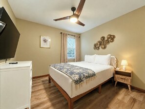 Une chambre à coucher comprenant un lit, une table de chevet, une lampe, un ventilateur de plafond, une fenêtre avec des rideaux et un téléviseur posé sur un meuble.