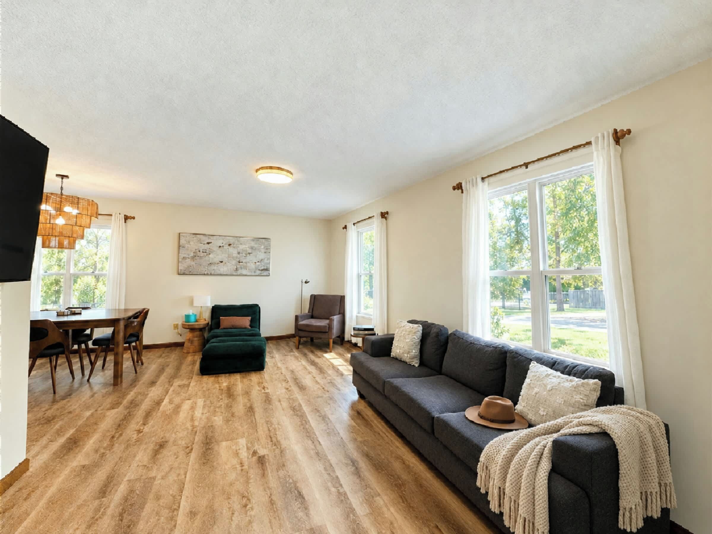 A spacious living room with a dark grey sofa, a wooden dining table with chairs, and large windows with curtains.