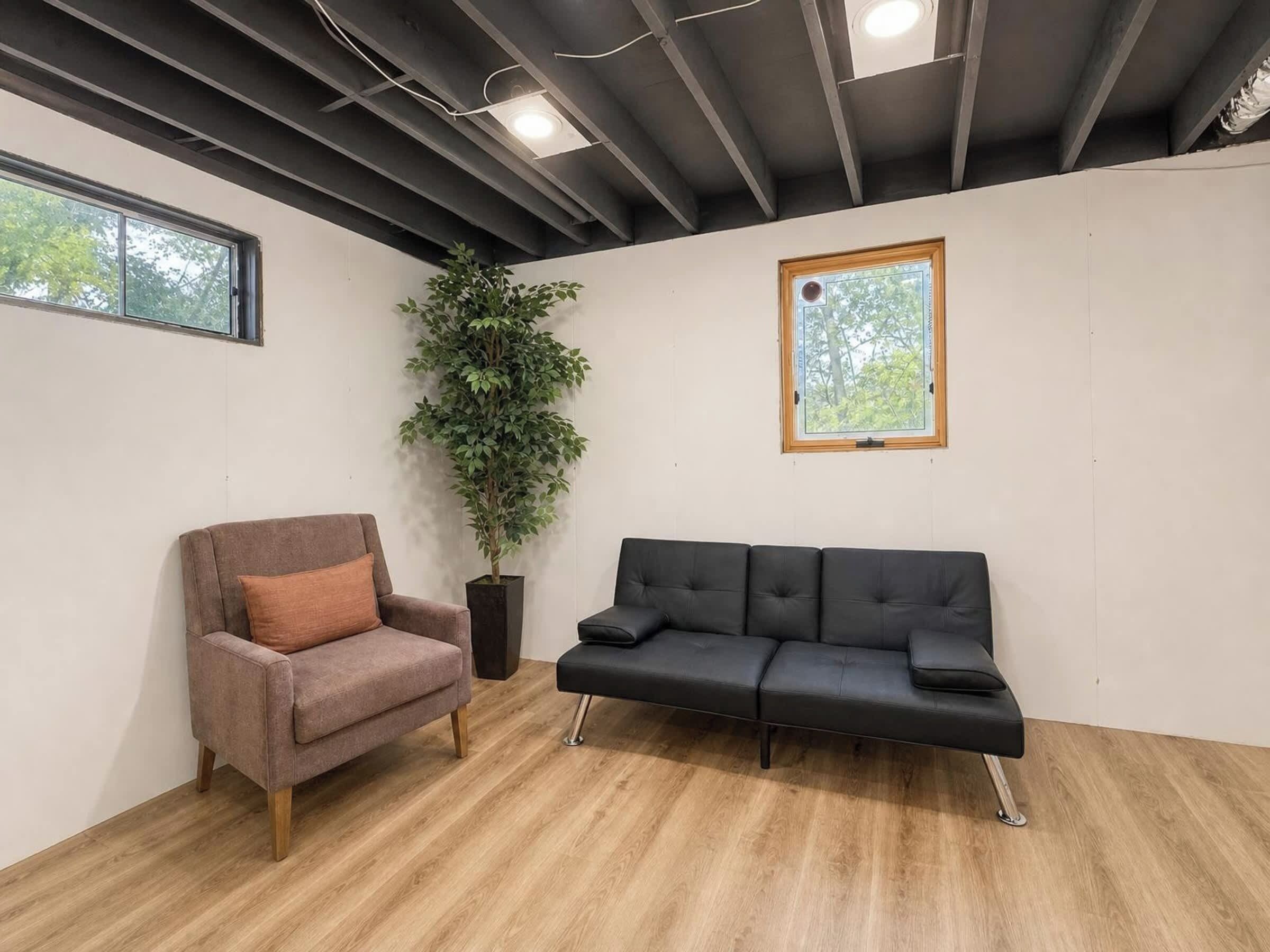 Una habitación con piso de madera, un sofá negro, un sillón marrón, una planta en maceta y una ventana con vista a árboles.