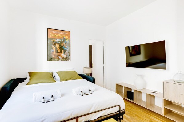 A modern hotel room with a neatly made bed, a flat-screen TV, a bedside table with a lamp, and a poster on the wall.