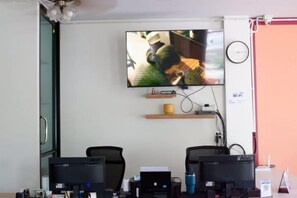 A room with a flat-screen TV mounted on the wall, two computer monitors on a desk, a clock, and a Wi-Fi sign. - Chill @ Phetchaburi (Phetchaburi)