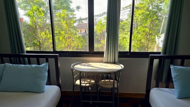 Una habitación con dos camas, una mesita redonda y un ventanal con vistas a árboles y edificios.