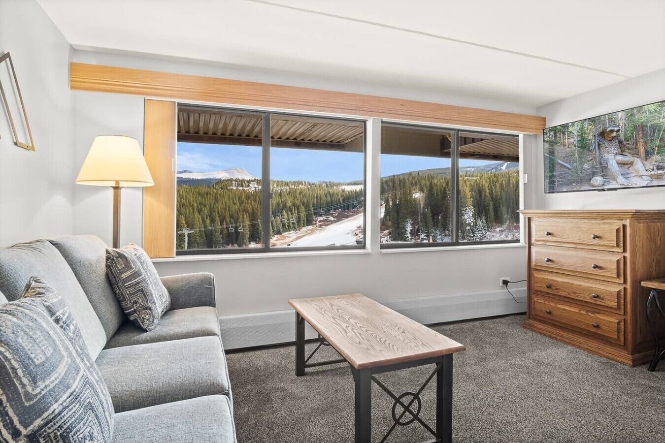 A room with a sofa, a table, a dresser, and a large window with a view of a snowy landscape and trees.