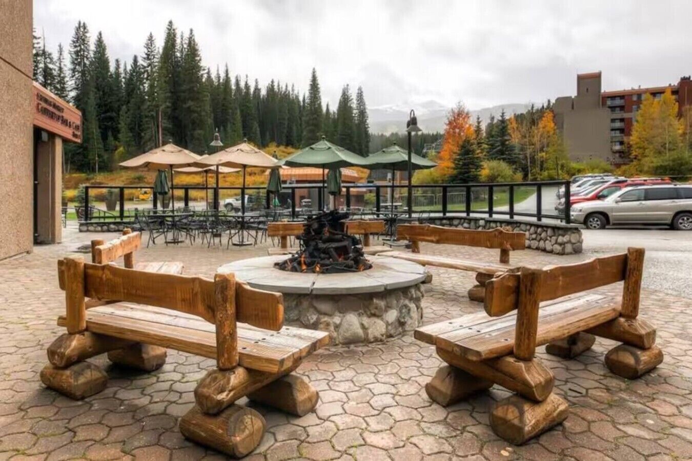 An outdoor seating area with wooden benches and a central fire pit, surrounded by umbrellas and a stone wall.