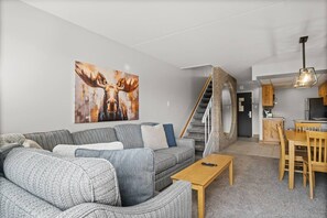 A modern living room with a grey sectional sofa, a wooden coffee table, and a staircase leading to a kitchen area.