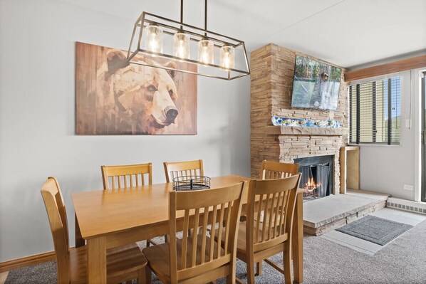 A dining area with a wooden table and chairs, a bear painting on the wall, and a stone fireplace.
