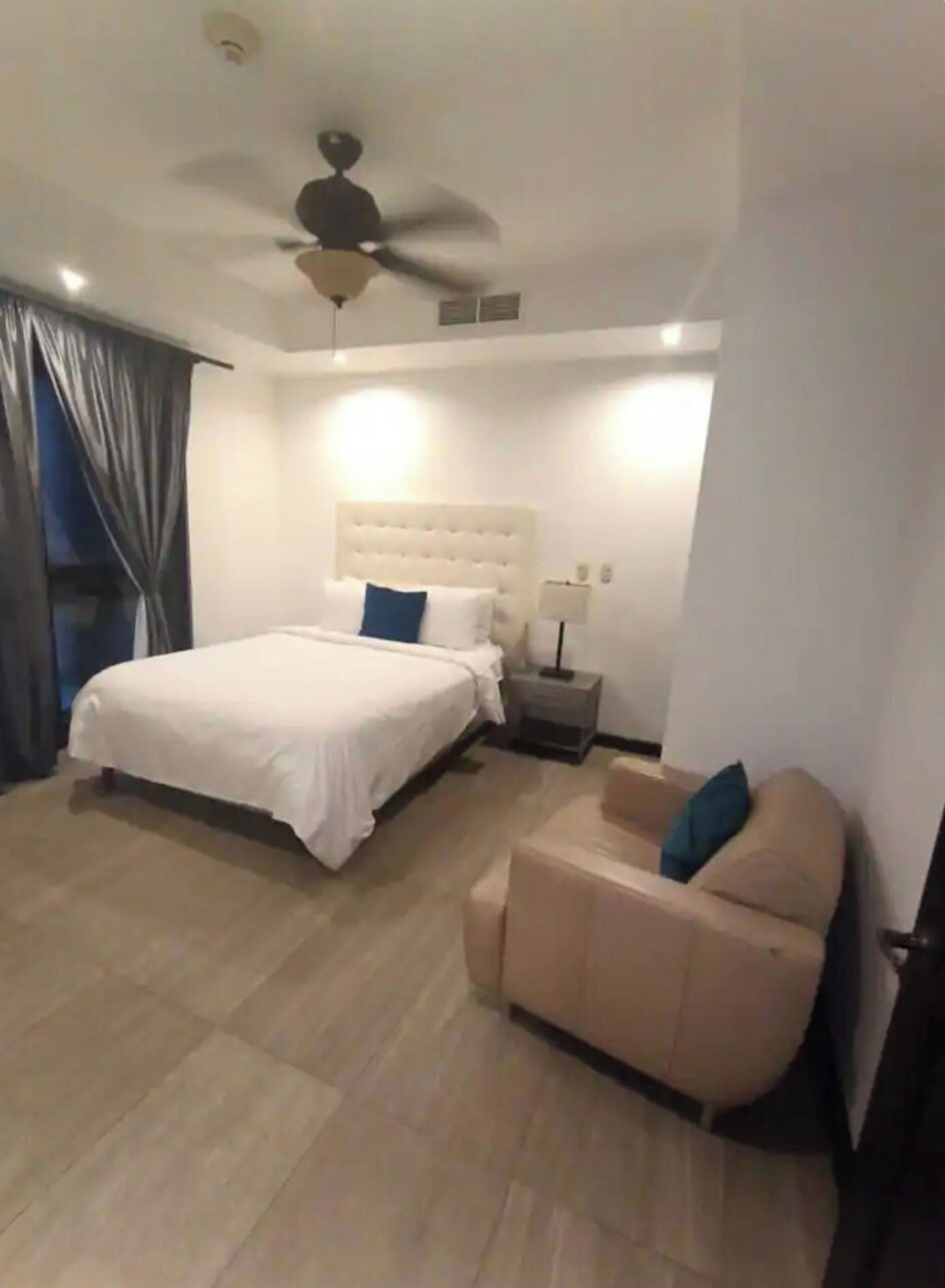 A bedroom with a bed, a ceiling fan, a nightstand, a lamp, and a sofa.