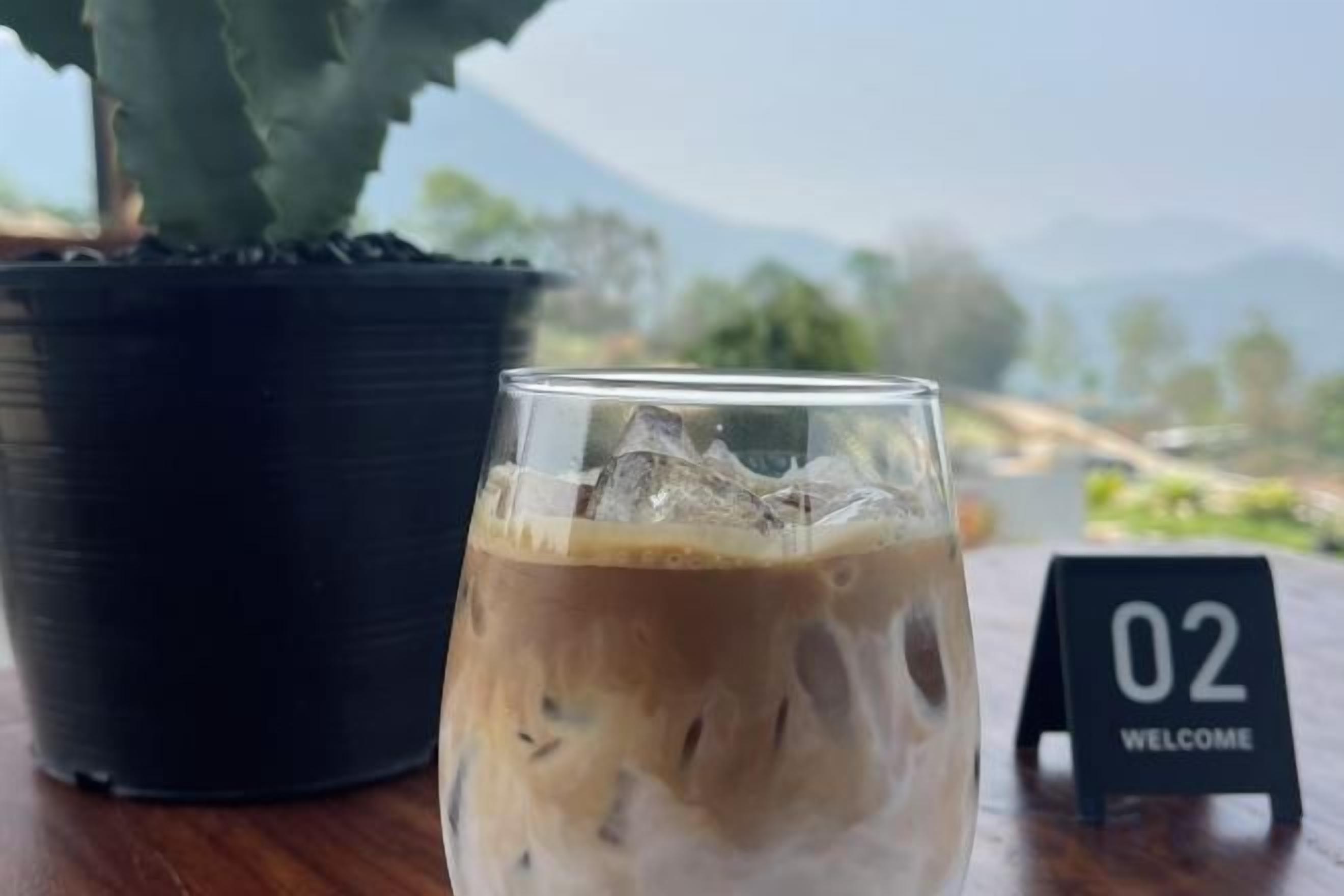 A glass of iced coffee on a table with a potted plant and a welcome sign.