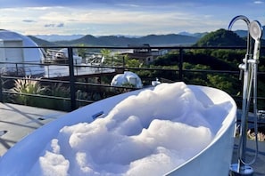 Eine Badewanne voller Blasen, ein Duschkopf aus Metall und Blick auf Berge und Grünland.