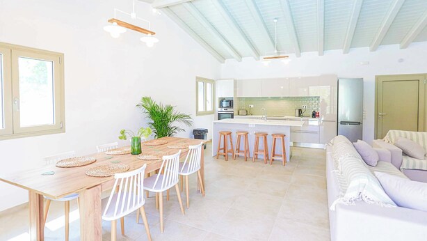 A spacious living area with a high ceiling, a dining table with chairs, a sofa, and a kitchen in the background. - Villa Jupeter (Corfu)