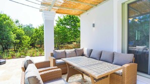 An outdoor patio with wicker furniture, a large table, and a glass door leading to a garden. - Villa Jupeter (Corfu)
