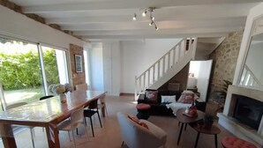 A spacious living area with a wooden dining table, a white armchair, and a sofa with red cushions. A staircase leads to an upper level, and a fireplace is visible on the right.