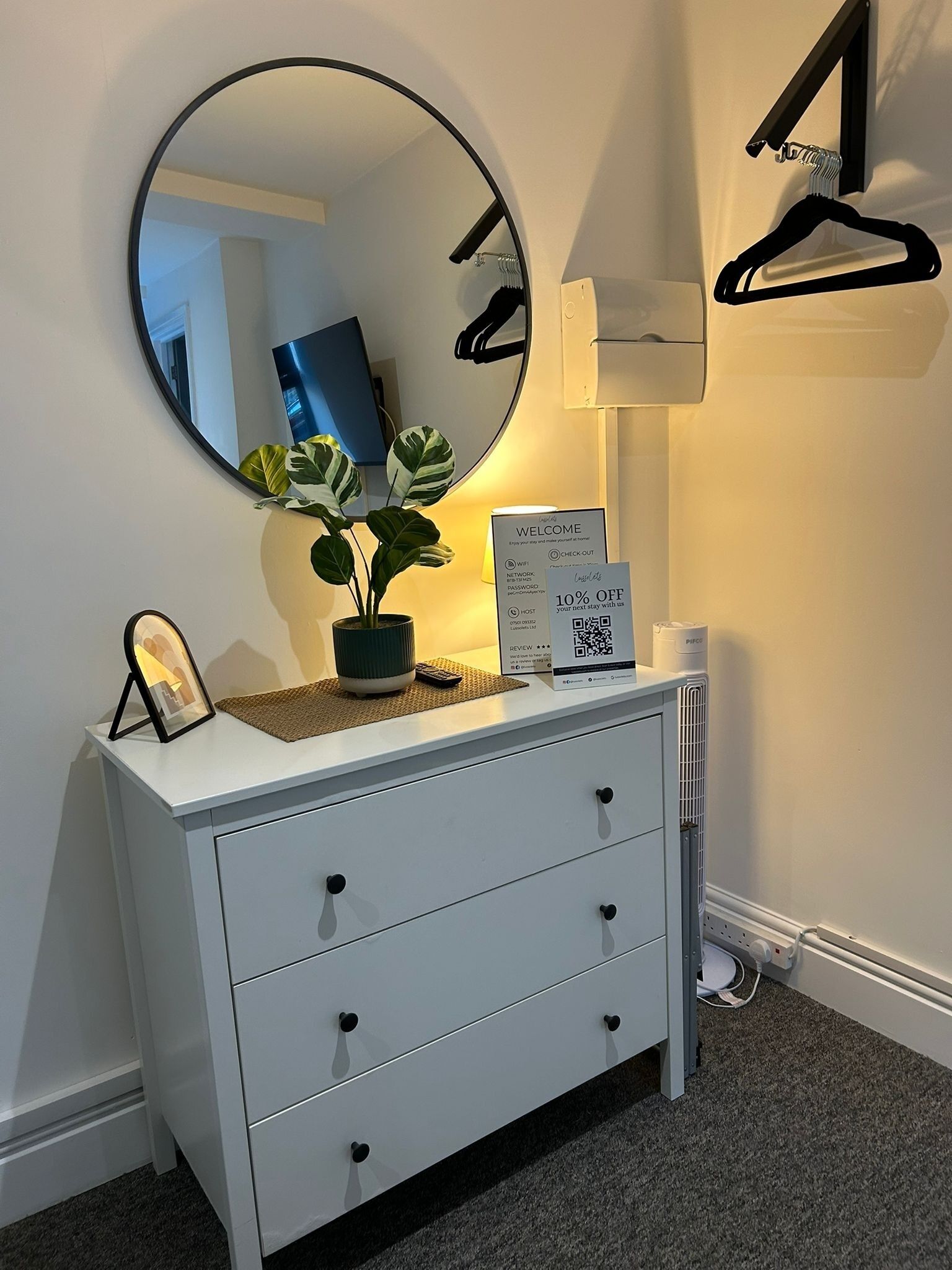 A white dresser with a round mirror, a small plant, a framed picture, and a welcome sign with a QR code.