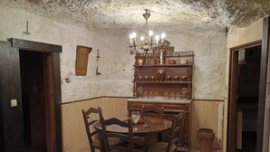 A rustic dining room with a wooden table and chairs, a chandelier, and shelves with pottery and glassware.