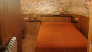 A small, rustic room with a single bed, a wooden bench, a small table with a radio, and a window with a curtain.