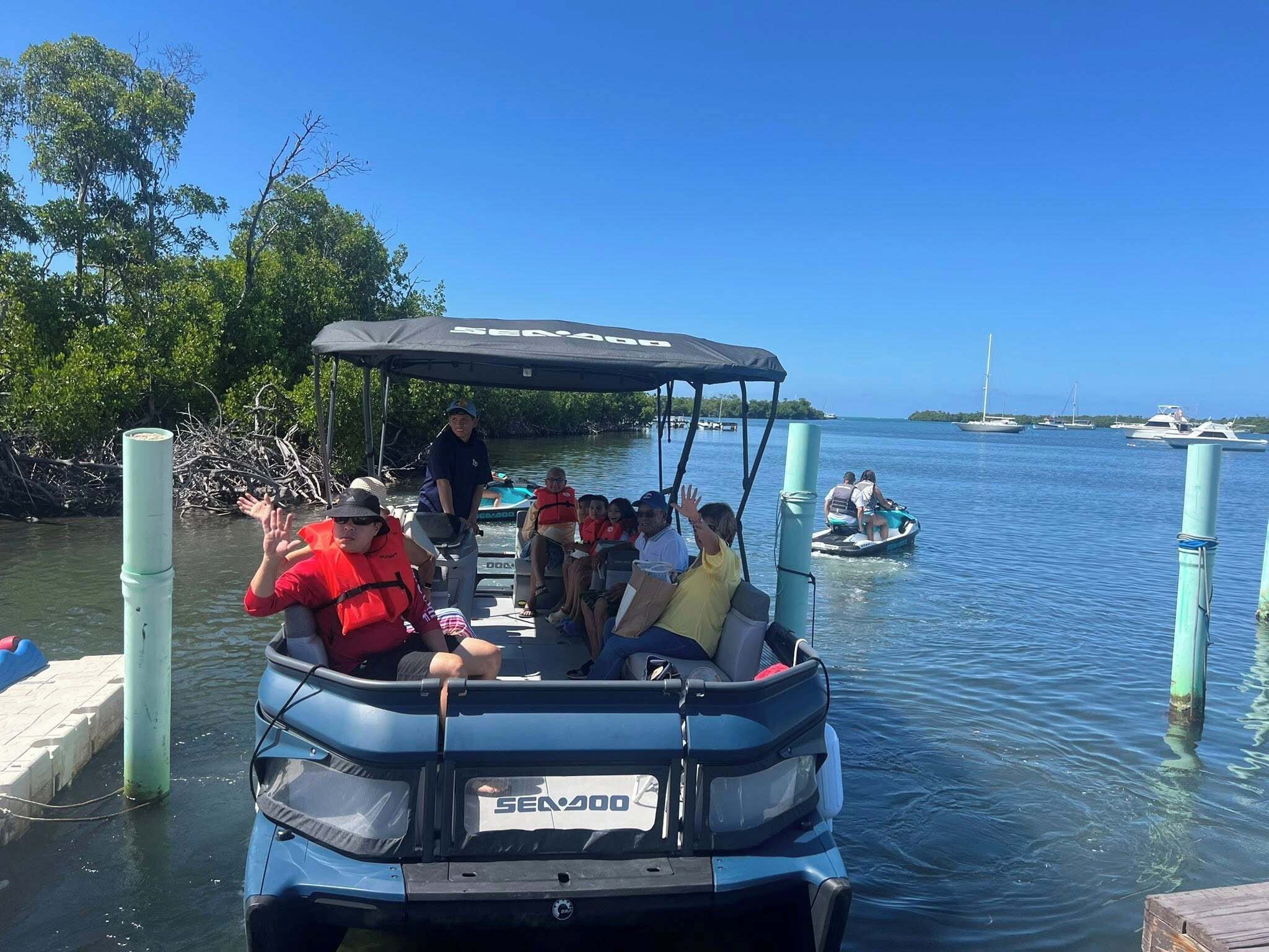 A Sea-Doo pontoon boat with passengers, docked at a marina with other boats and a clear blue sky.