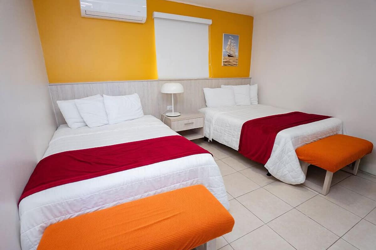 A hotel room with two beds, each with a red and white bedspread, a nightstand with a lamp, and a window with a view of the sea.