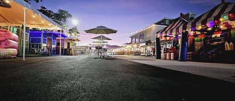 A street at dusk with outdoor seating and umbrellas, shops with colorful facades, and a clear sky.