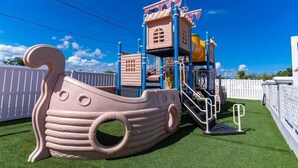 Une structure de jeux pour enfants en forme de bateau, dotée d’un toboggan, entourée de clôtures blanches et d’une pelouse artificielle verte.