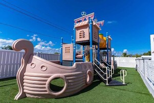 Una estructura de juego para niños con forma de barco, que incluye un tobogán y está rodeada de vallas blancas y césped artificial verde.