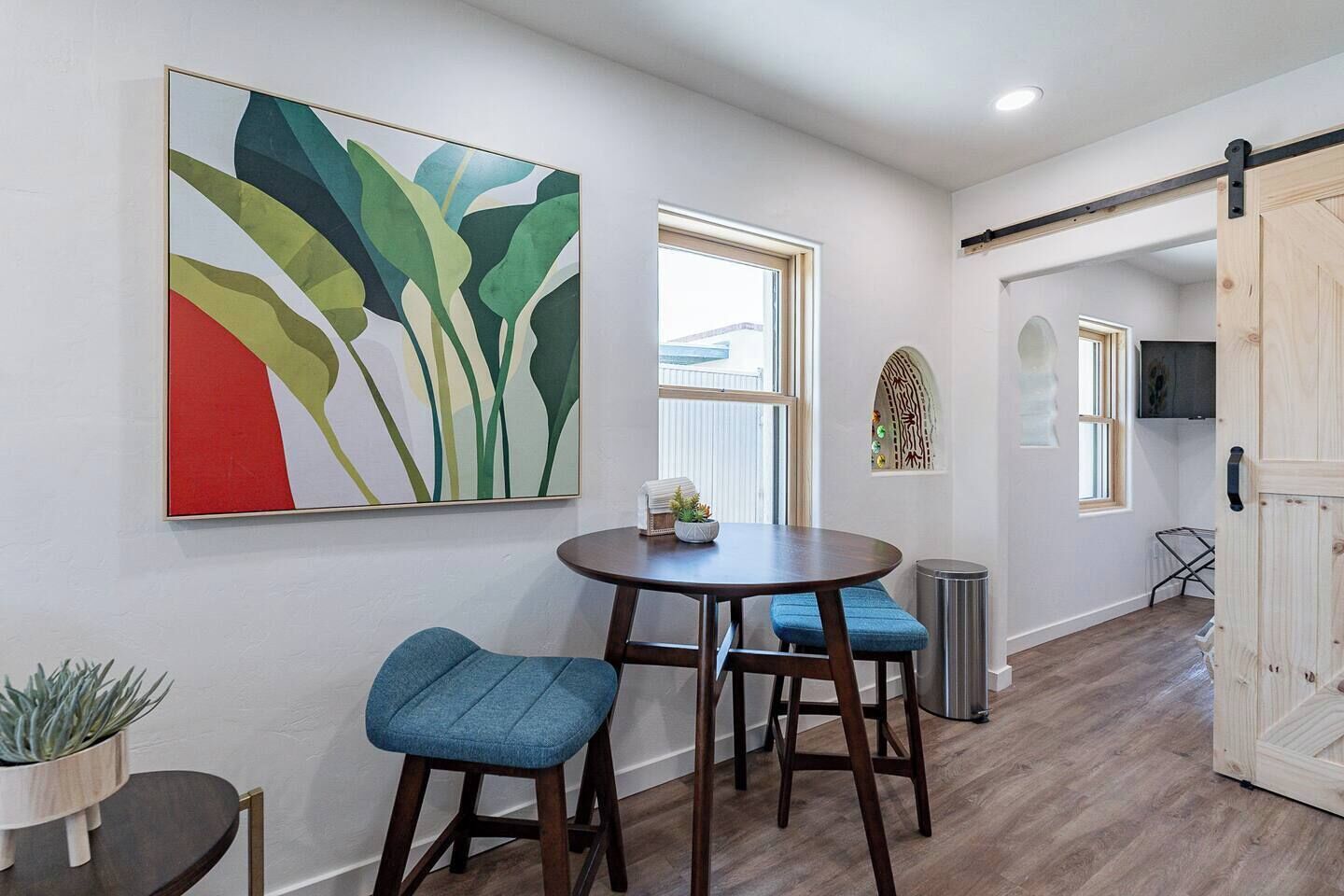 A modern dining area with a round table and blue upholstered chairs, a large abstract painting on the wall, and a sliding barn-style door.