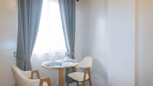 Une petite pièce avec une table ronde, deux chaises, une fenêtre avec des rideaux et un interrupteur sur le mur.