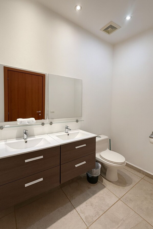 A modern bathroom with a double sink vanity, a toilet, and a trash bin.