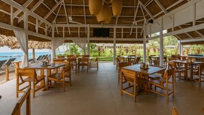 A covered outdoor dining area with wooden tables and chairs, a thatched roof, and a view of the ocean. - Barmel Villa Rooms (Roatan)