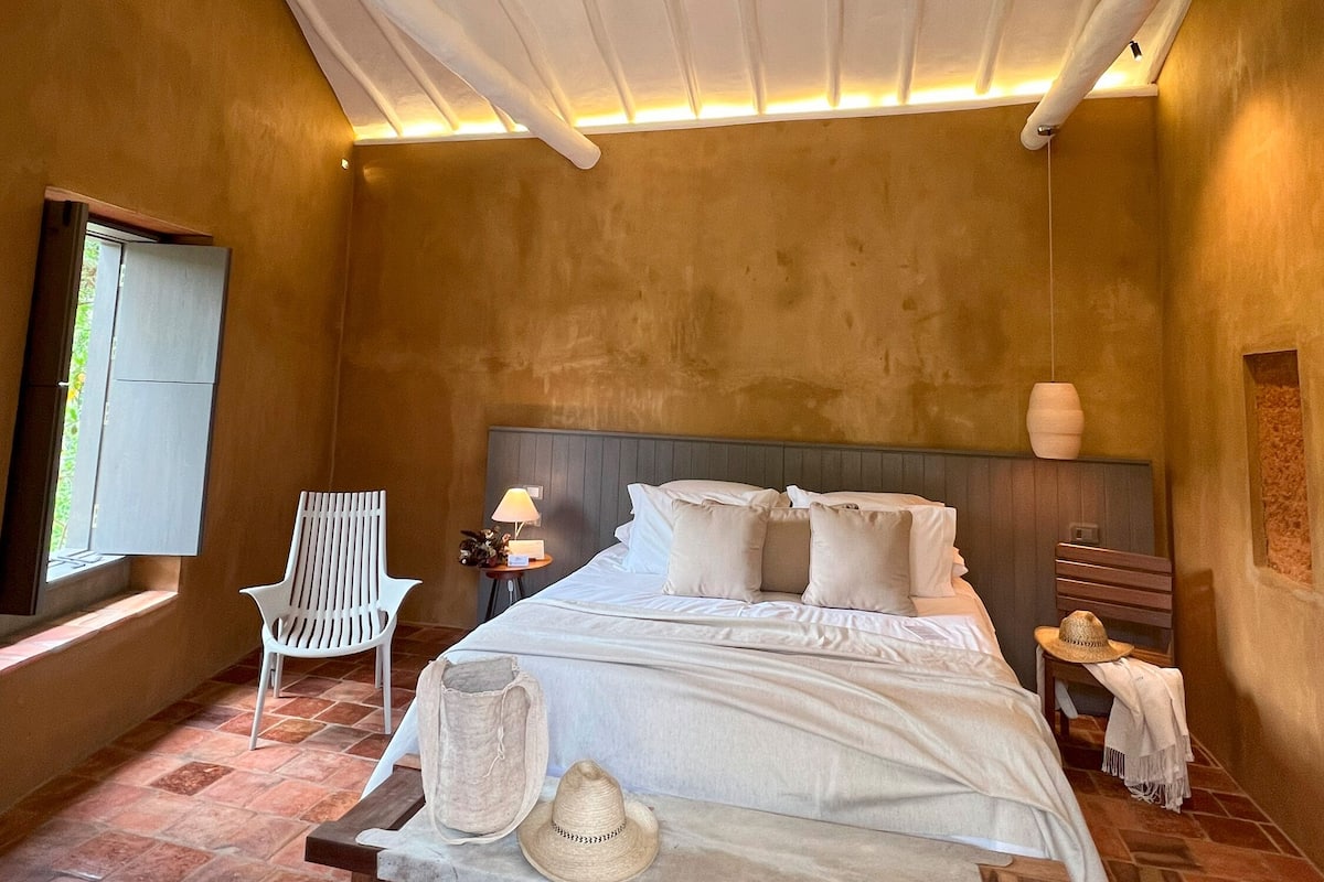 Une chambre à coucher avec un lit, une chaise, une table de chevet, une lampe et une fenêtre.
