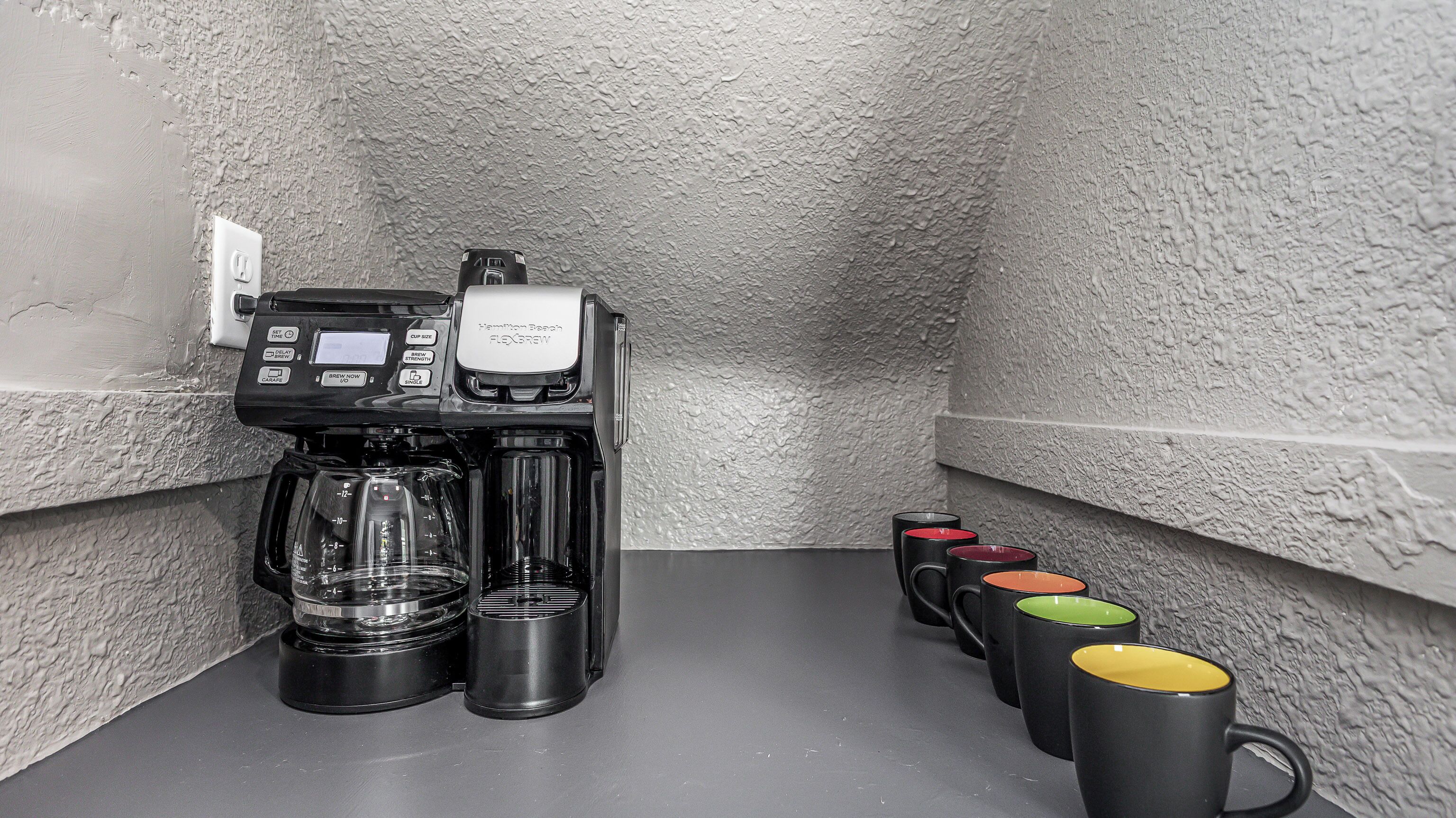 Una cafetera con depósito de agua y bandeja de goteo, un enchufe, y una fila de tazas de colores en una pared con textura.