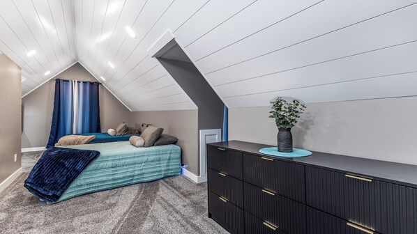 Une chambre avec un plafond en pente, un lit recouvert d’une couverture bleue, une commode et une plante en pot.