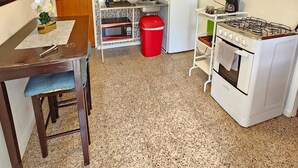Une cuisine compacte équipée d’une cuisinière à gaz, d’un four à micro-ondes et d’un petit réfrigérateur. On y trouve une table à manger avec une chaise, une poubelle rouge et une étagère contenant des ustensiles de cuisine.